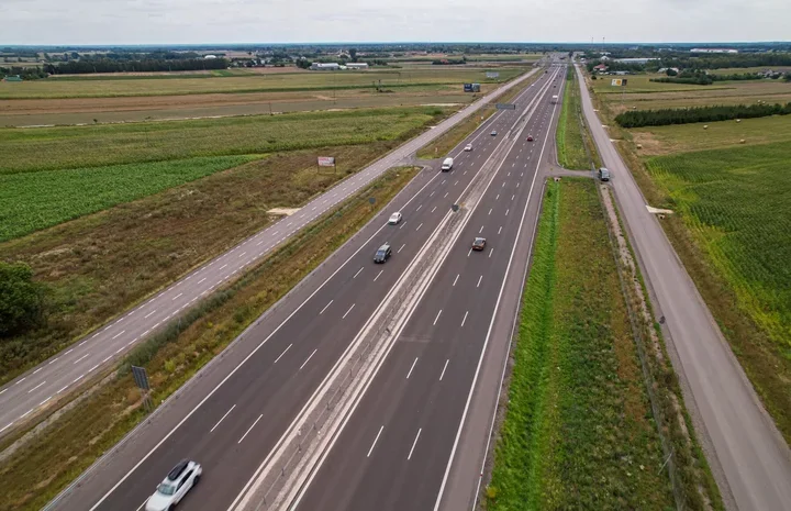 Wielopasmowa autostrada biegnie przez płaskie, zielone tereny rolnicze, po której poruszają się pojazdy.