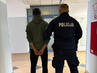Funkcjonariusz policji stoi obok zatrzymanego mężczyzny w kajdankach w korytarzu budynku.