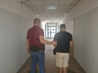 Funkcjonariusz w czerwonej koszulce prowadzi zatrzymanego mężczyznę w czarnej koszulce i beżowych szortach przez korytarz budynku urzędowego.