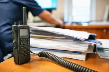 Radiotelefon leży na biurku obok stosów dokumentów, w tle siedzi osoba w ciemnej koszuli przy biurku.