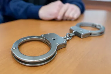 Metalowe kajdanki leżą na drewnianym stole, w tle widoczne splecione dłonie osoby w granatowej bluzie.