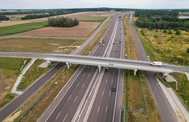 Widok z lotu ptaka na wielopasmową autostradę z wiaduktem drogowym, otoczoną polami i lasami, z pojazdami poruszającymi się po jezdniach.